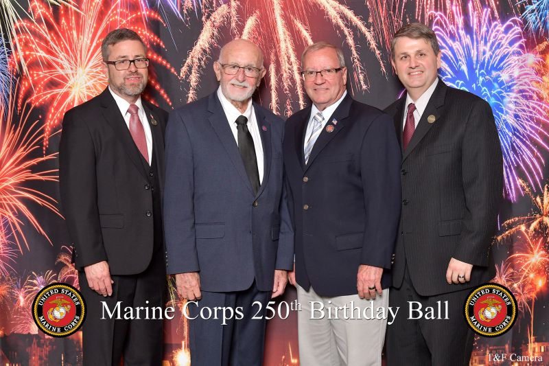 Four men pose at the Marine Corps 250th Birthday Ball, with one holding a framed certificate in front of a fireworks-themed backdrop