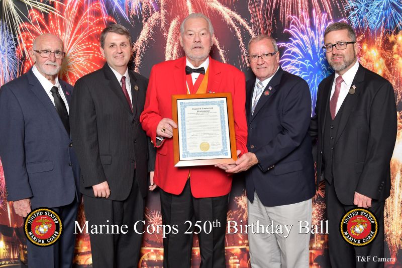 Five men pose at the Marine Corps 250th Birthday Ball, with one holding a framed certificate in front of a fireworks-themed backdrop