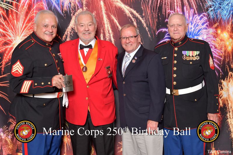 Four men pose at the Marine Corps 250th Birthday Ball, with one holding a framed certificate in front of a fireworks-themed backdrop